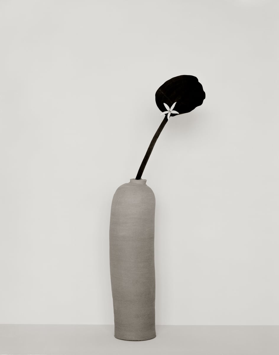 A dark lotus bud with a tiny white star-shaped flower, leaning from a tall grey stoneware vessel, in black and white